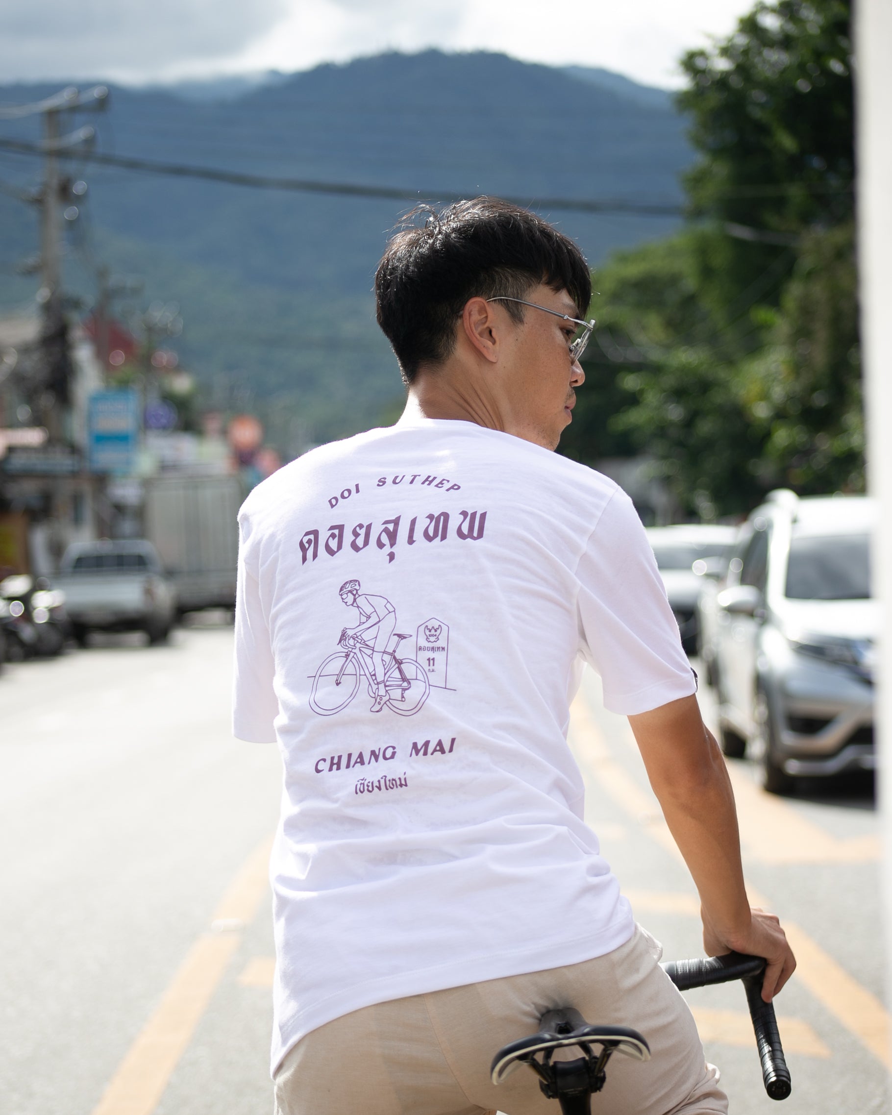 A man wearing a white t-shirt with a purple graphic print on the back, riding a bicycle on a street with mountains in the background.