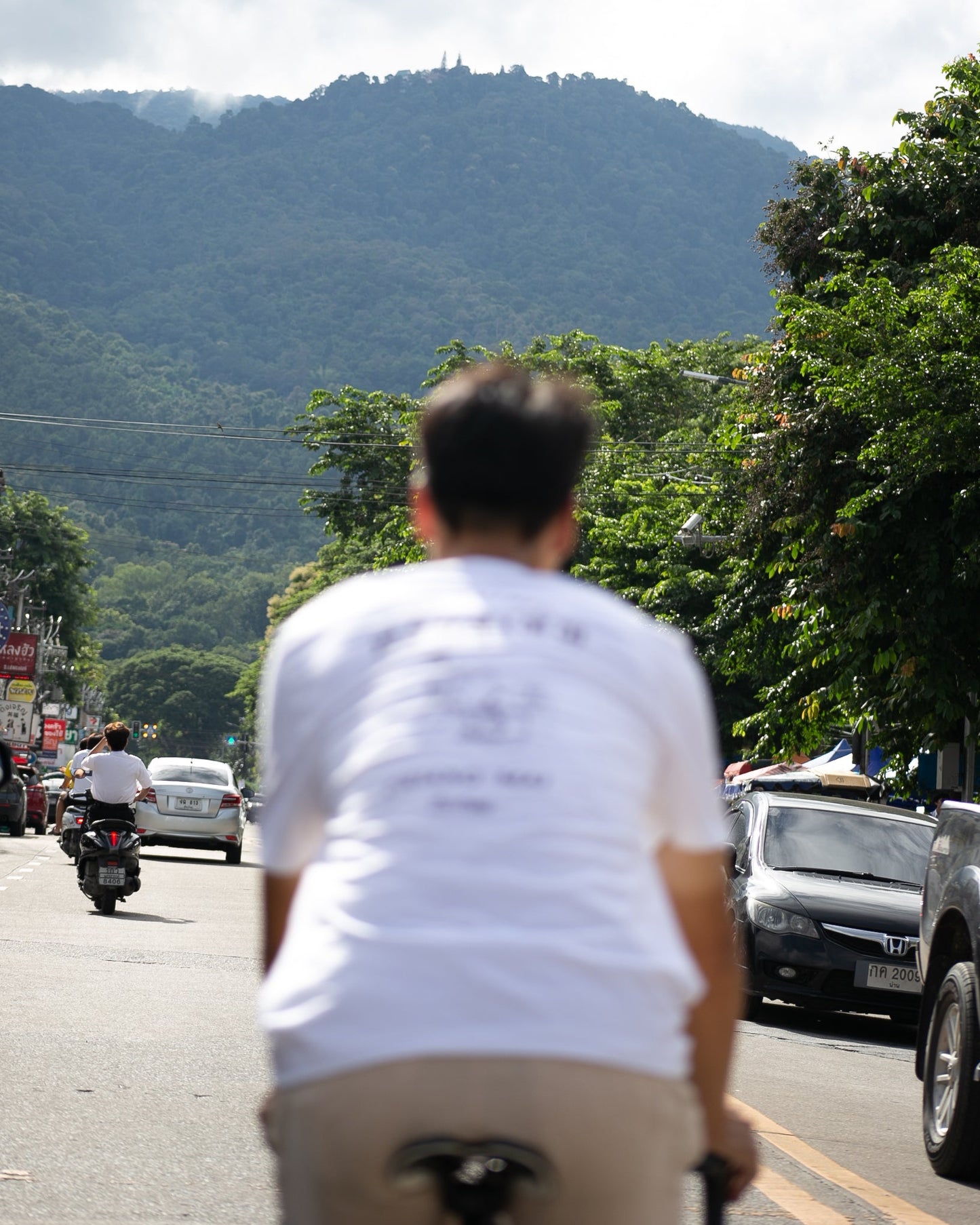 A person seen from behind wearing a white t-shirt and beige pants, riding a bicycle on a busy road with cars, motorcycles, and a lush green mountain in the background.