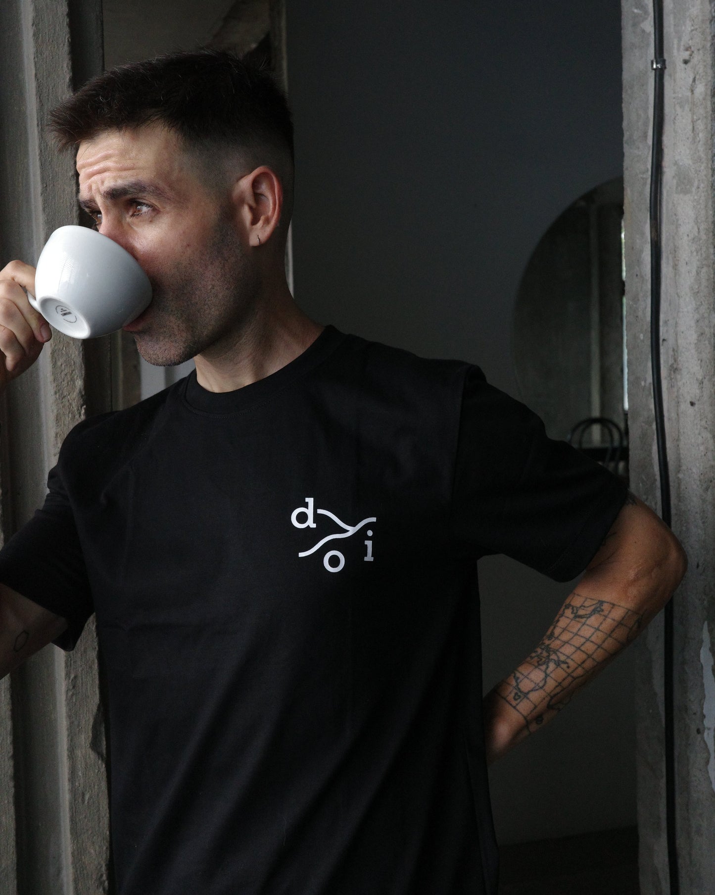 A man wearing a black t-shirt with a white stylized 'doi' logo on the chest, drinking from a white mug in a dimly lit, rustic interior.