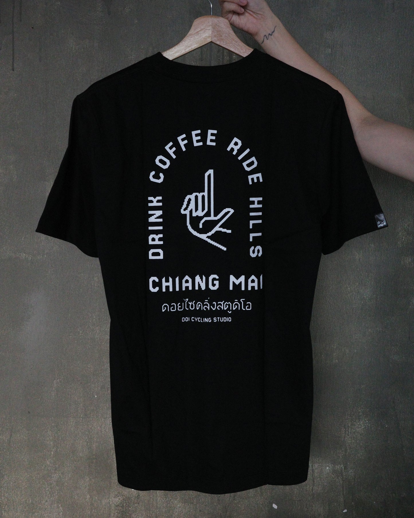 A black short-sleeve t-shirt with a white graphic print on the back, hanging on a wooden hanger held by a person's hand against a dark textured wall.