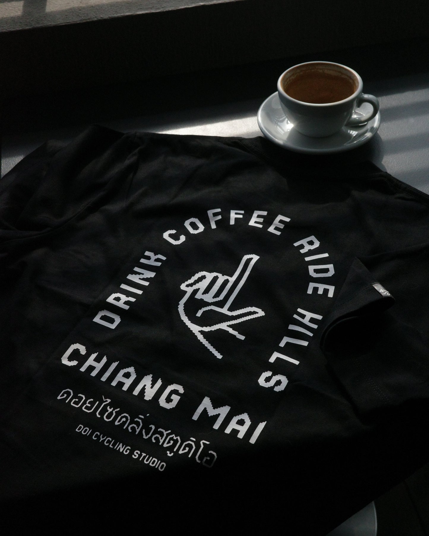 A black t-shirt with a white graphic print reading 'DRINK COFFEE RIDE HILLS CHIANG MAI' and 'Doi Cycling Studio' is laid out on a dark table next to a cup of coffee on a saucer.