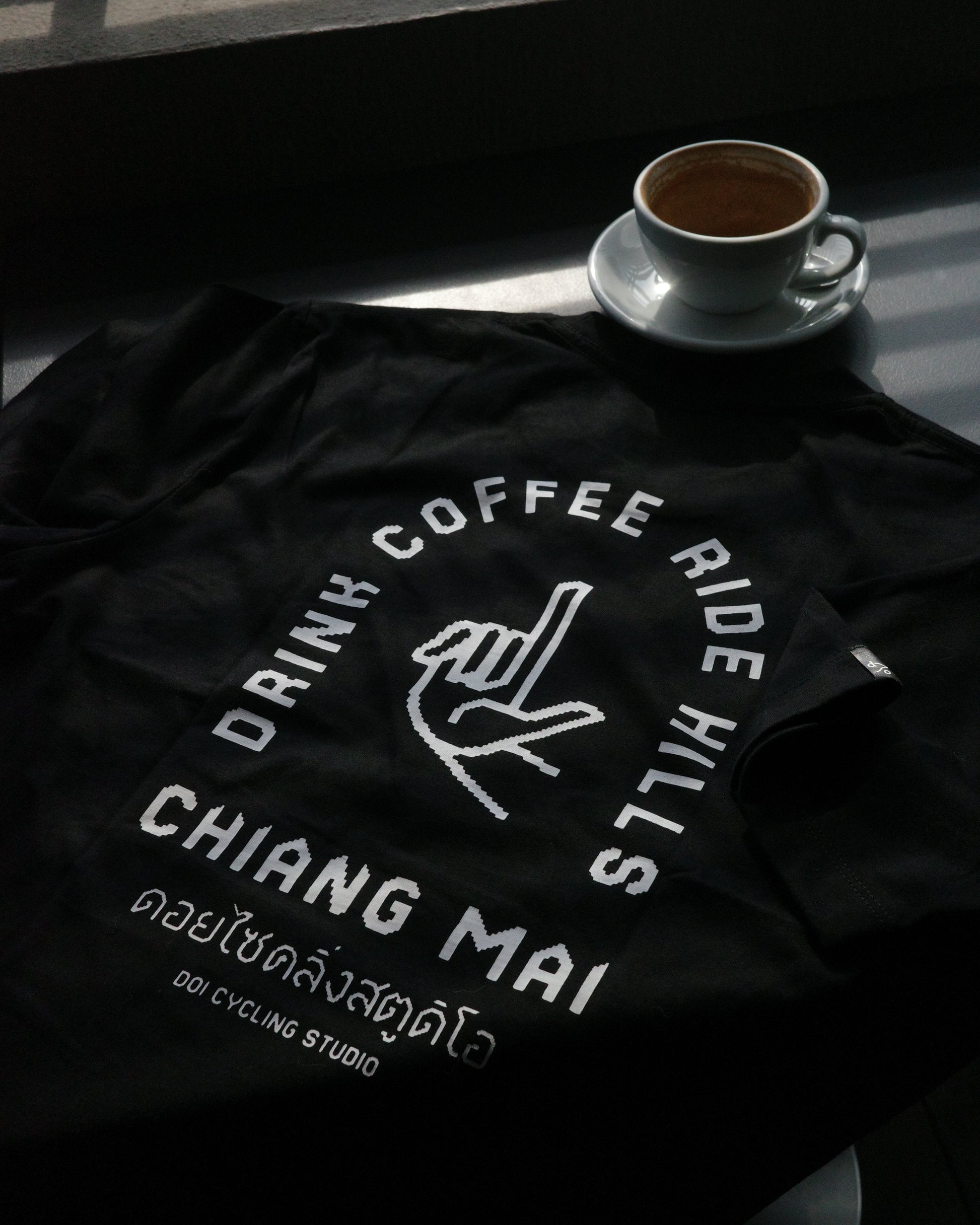 A black t-shirt with a white graphic print reading 'DRINK COFFEE RIDE HILLS CHIANG MAI' and 'Doi Cycling Studio' is laid out on a dark table next to a cup of coffee on a saucer.