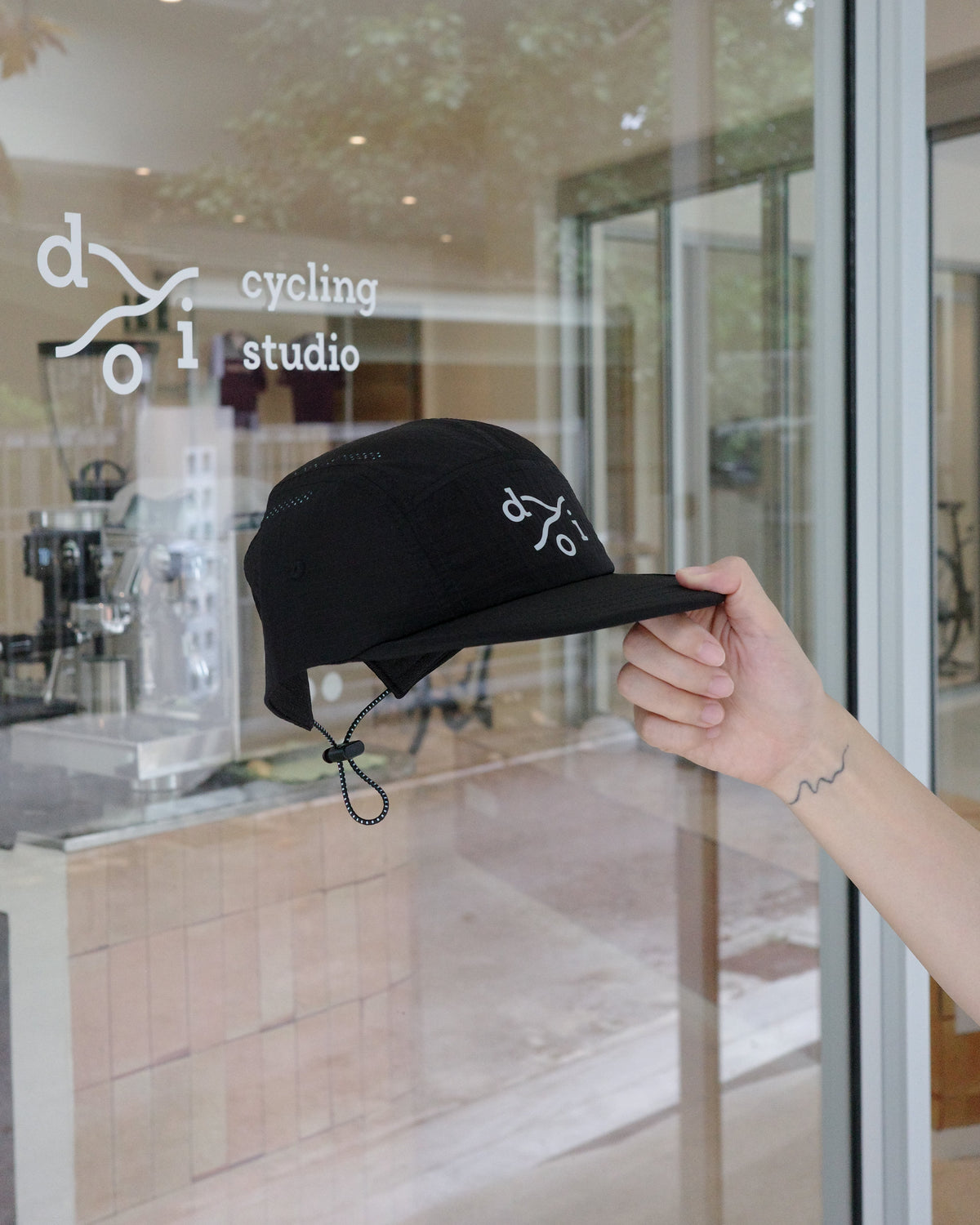 A black cycling cap with reflective logo details is held by a hand in front of a glass window of a 'doi cycling studio'.