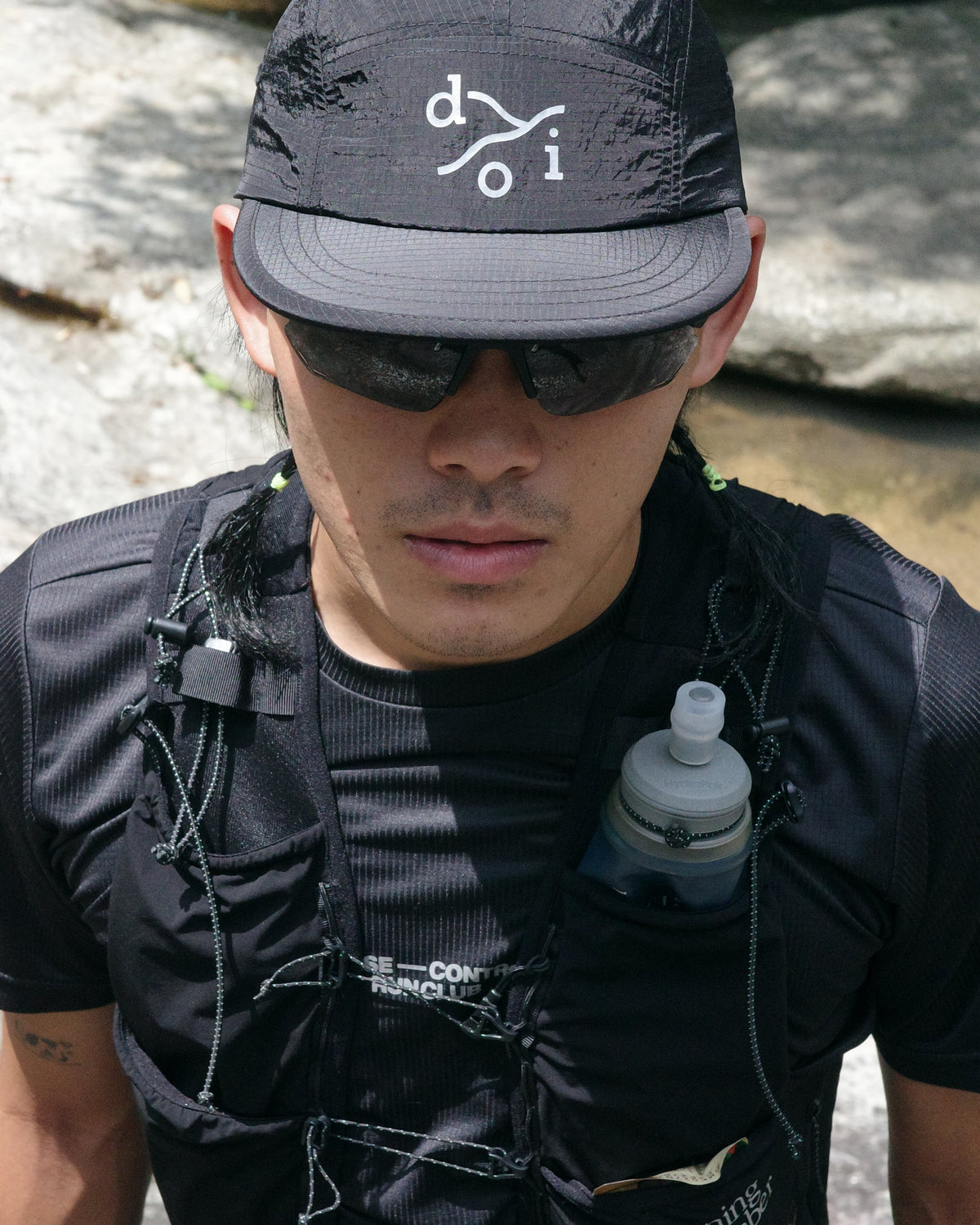 A close-up of a man outdoors wearing a black running cap with a reflective logo, black sunglasses, a black running shirt, and a black hydration vest with a water bottle in the pocket. The background shows rocks and a stream.