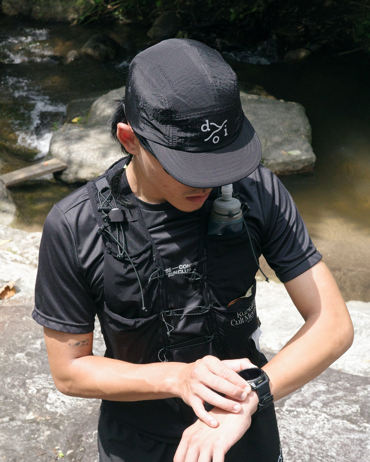 A man in black athletic gear, including a black cap with a white 'doi' logo, a black hydration vest, and black shorts, checks the time on a smartwatch outdoors near a stream.