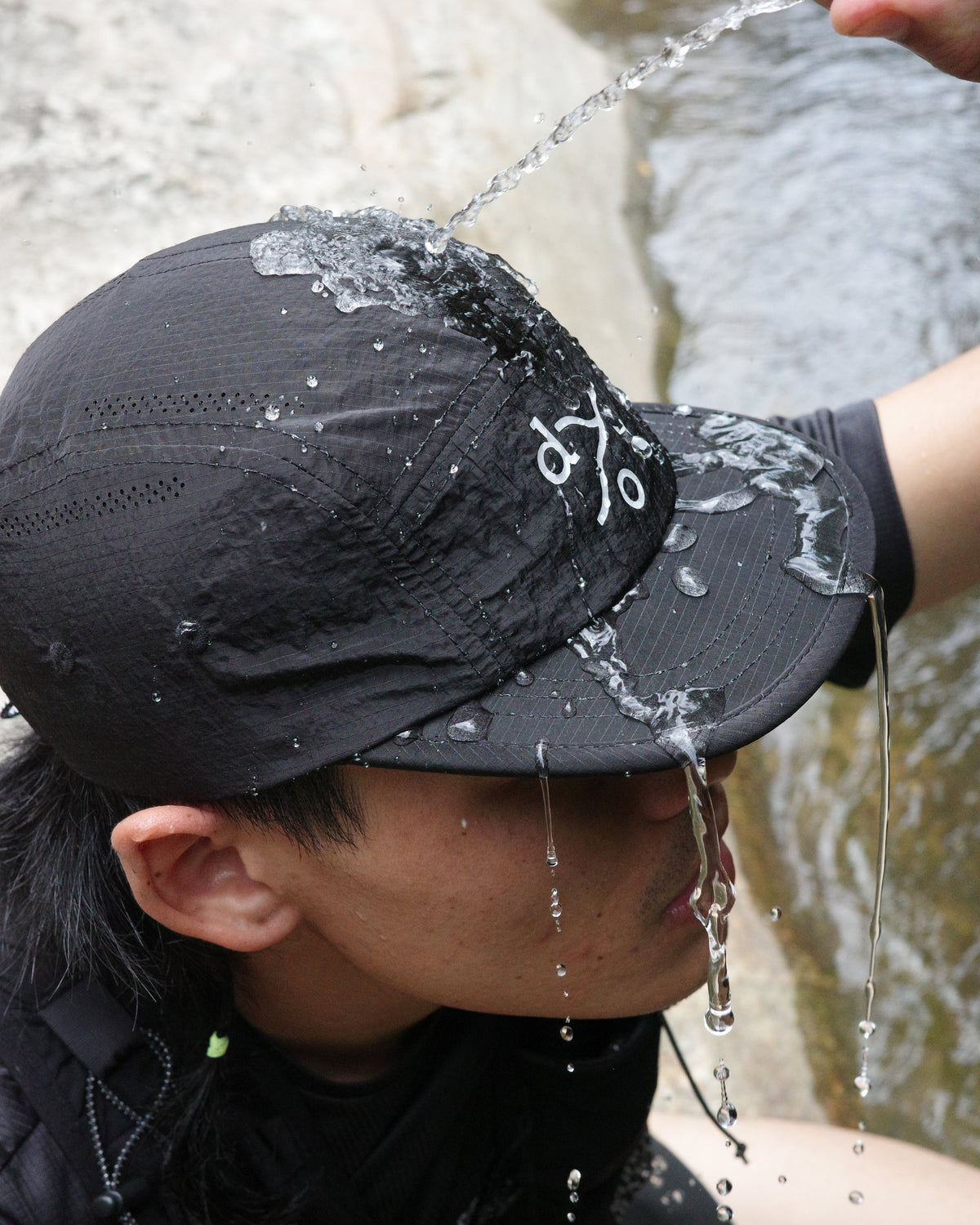 A close-up lifestyle shot of a person outdoors in a stream, wearing a black athletic cap with reflective text and pouring water from a bottle onto their head.