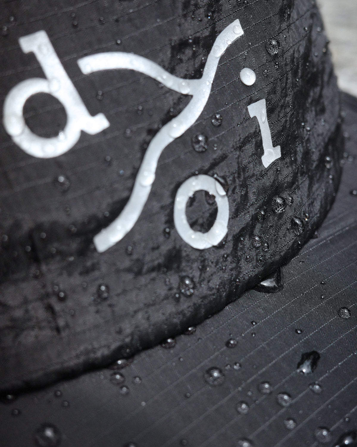 Close-up of a black cap covered in water droplets, with a reflective stylized logo across the front.